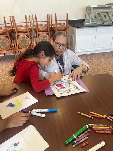 A young child colouring at a table alongside a resident of a memory care centre