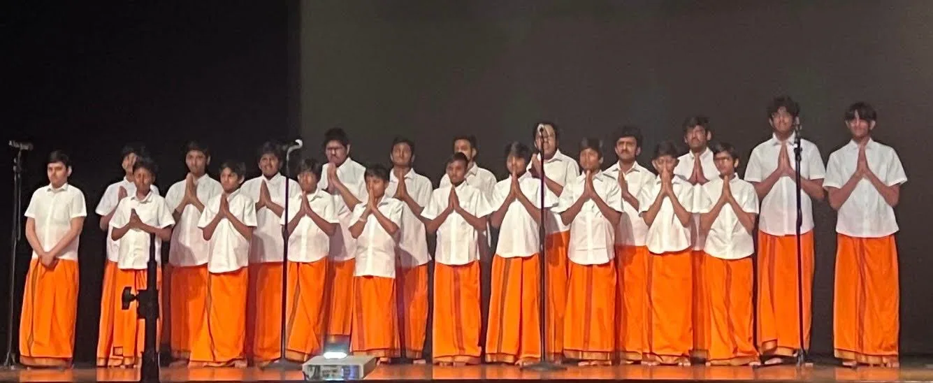 Bal Vikas students in white and saffron dhotis standing in a line on stage with hands folded, chanting